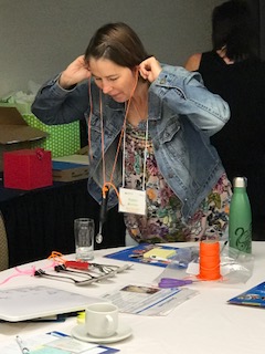 An educator has a metal spoon tied with yarn and the ends of the yarn are wound around her index fingers. Her fingers are in her ears as she taps the metal spoon against an empty drinking glass 