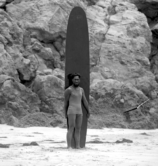 Surfer and Olympic swimmer Duke Kahanamoku standing on a beach with a surfboard in Los Angeles, California, circa 1920 