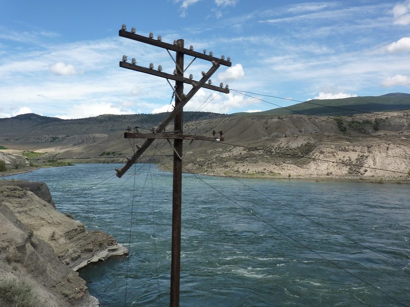 Shown is a colour photograph of a wooden pole connected to many wires, next to a river.