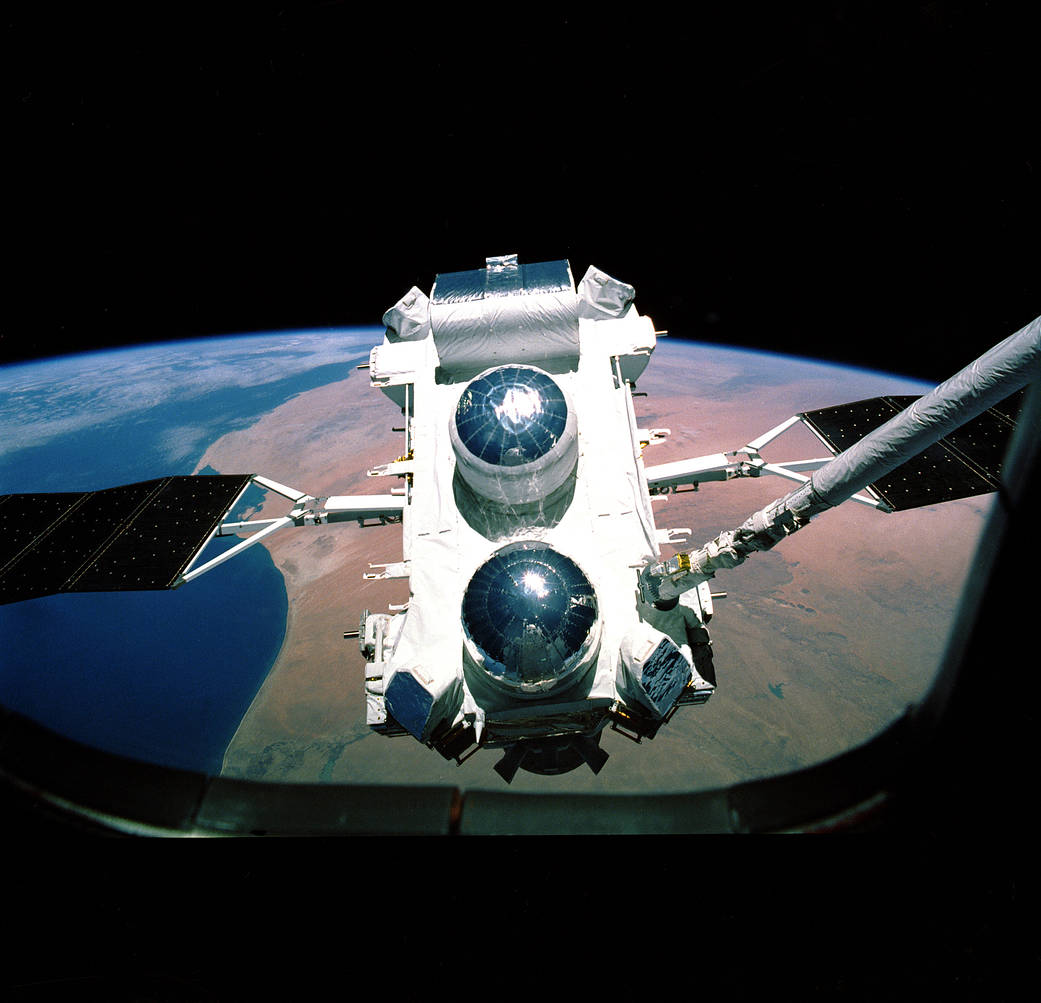 Shown is a flat, white rectangular structure with two large silver domes, floating above Earth.