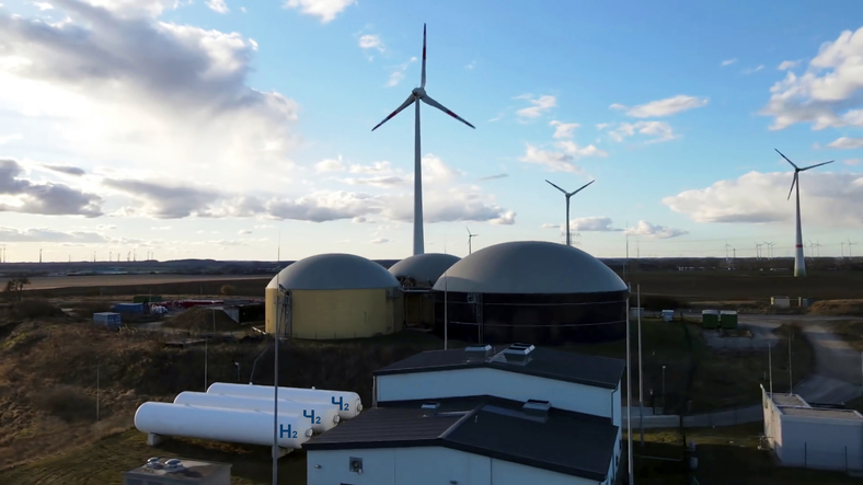 Shown is a colour photograph of buildings, wind turbines, and three tanks marked H2.
