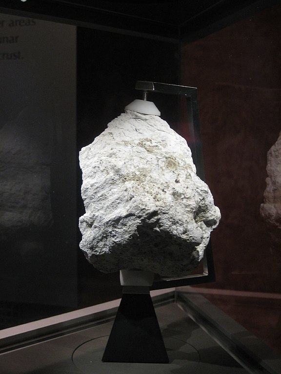 Shown is a colour photograph of a pale grey rock on a stand in a dark museum case.