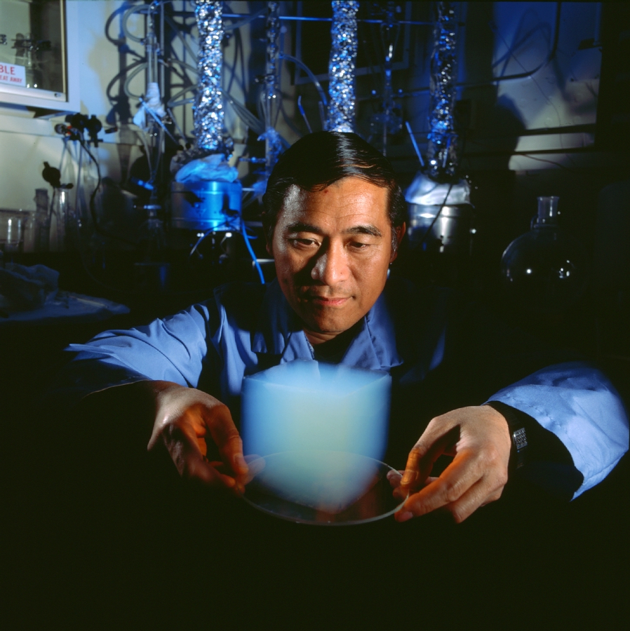 Shown is a colour photograph of a JPL scientist holding a translucent cube of aerogel on a glass plate, with arms bent at the elbows.