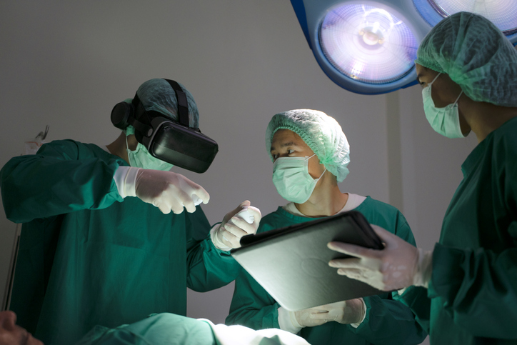 Shown is a colour photograph of three people in surgical scrubs, one is wearing VR goggles and another is holding a tablet.