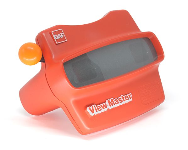 Shown is a colour photograph of a red plastic object with eyepieces, an orange lever, and a grey panel.