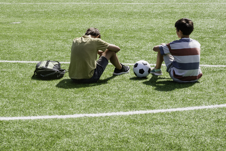 Shown is a colour photograph of two young people sitting on a soccer field with a ball between them.