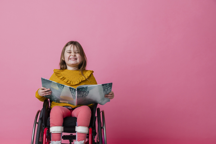 Shown is a colour photograph of a smiling young child seated in a wheelchair, holding an open book over their lap.