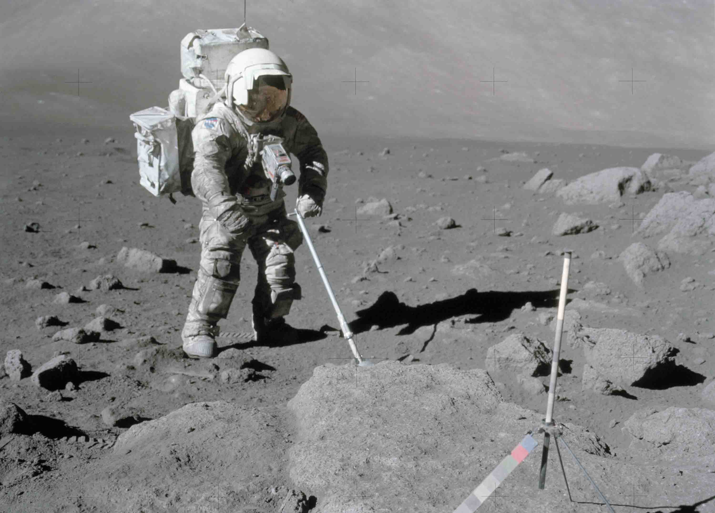 Shown is a black and white photograph of a person dressed in a space suit, extending a rod-like tool into the ground on the Moon.