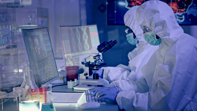 Shown is a colour photograph of two people in full protective gear, working at a lab bench.
