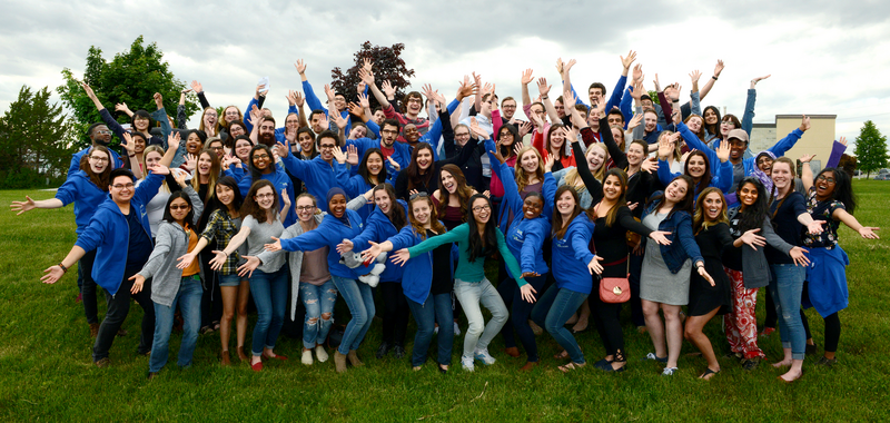A large group of Let's Talk Science volunteers pose together outside, smiling, with their arms stretched out