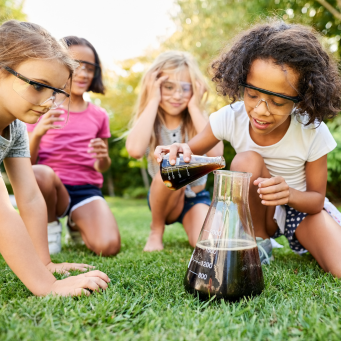Shot of little girls experimenting in garden, with a girl adding cola in a big glass flask