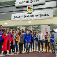 Large group of volunteers in wizarding costumes posing in front of banner saying "Let's Talk Science School of Witchcraft and Wizardry"