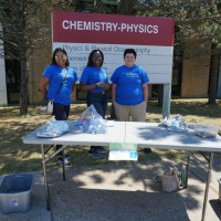 three Let's Talk Science Volunteers standing at an activity table in front of a Chemistry & Physics sign