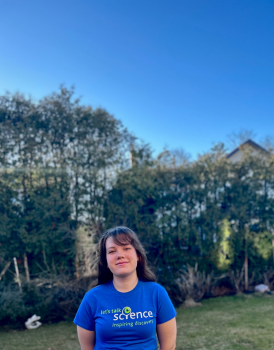 Julia stands in a field against the backdrop of green foliage and bright blue skies. She is wearing a blue Let's Talk Science t-shirt, with her brown hair falling over her shoulders as she smiles at the camera.