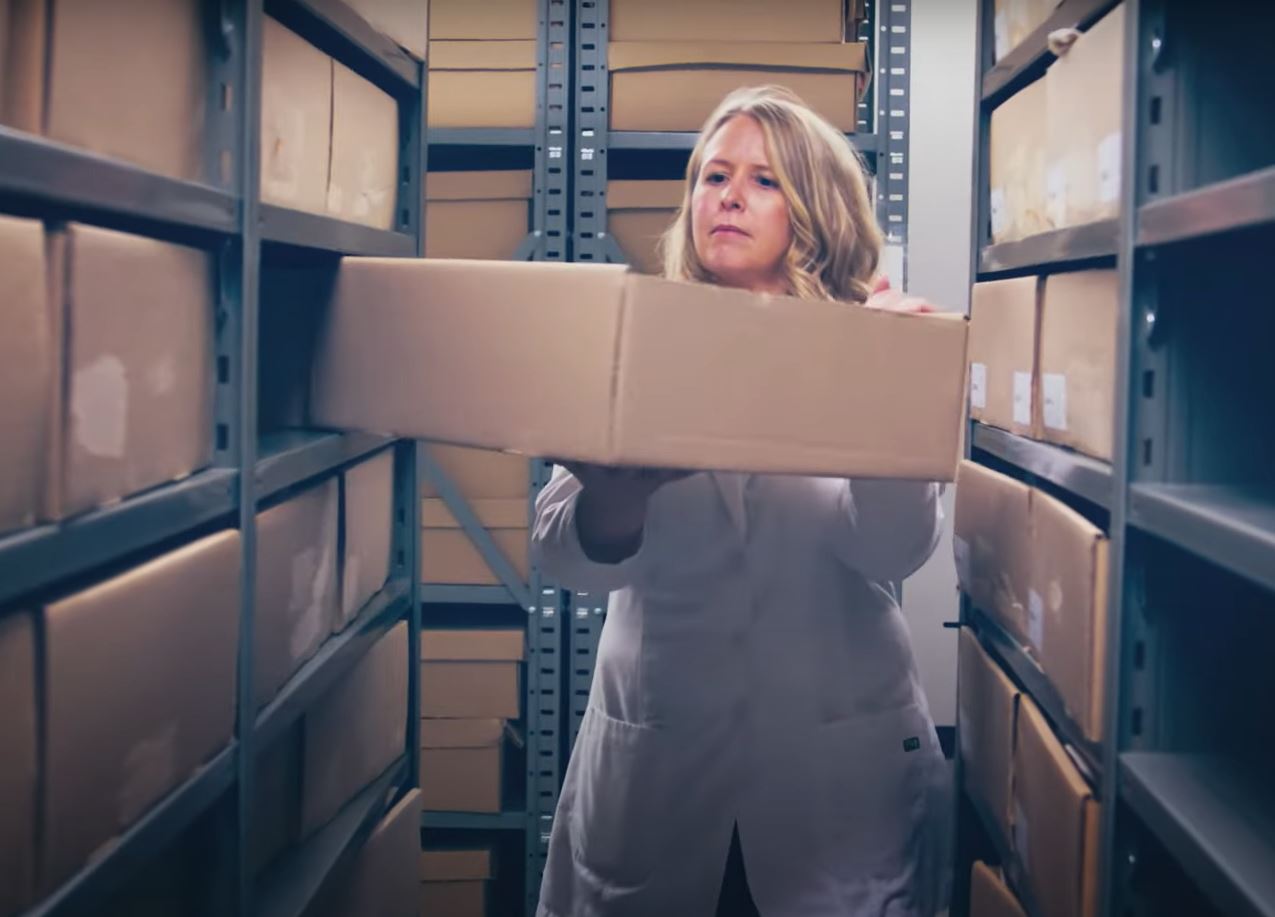 Zoe Ehlert wearing white lab coat, placing a cardboard box into a shelving unit