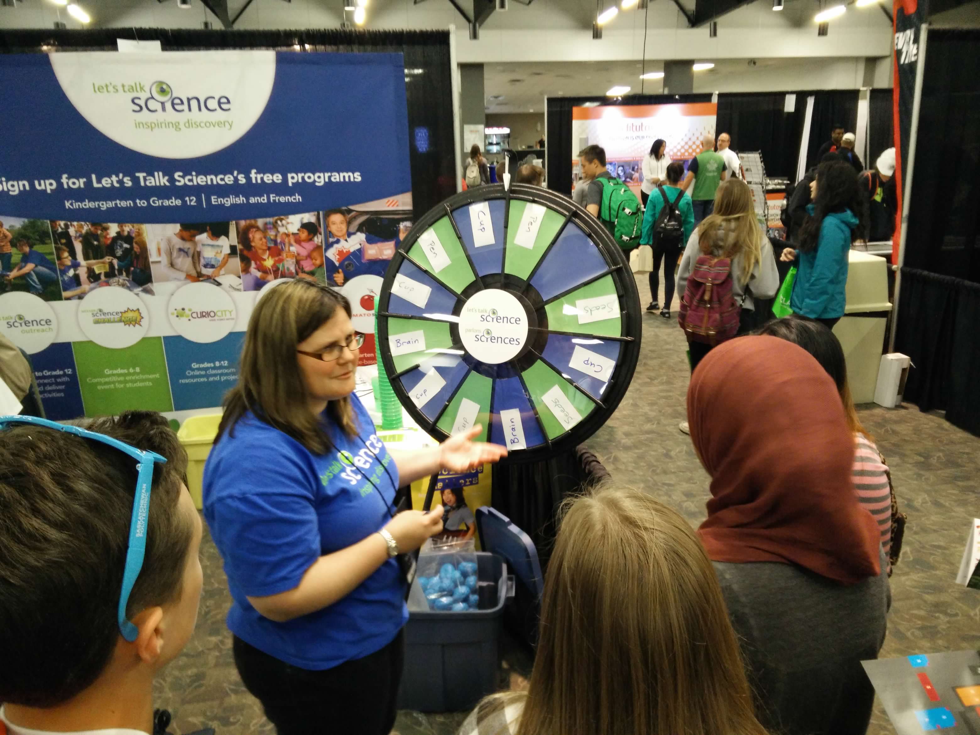 Glenda at a conference in a blue Let's Talk Science shirt talking to students in front of a blue and green prize wheel