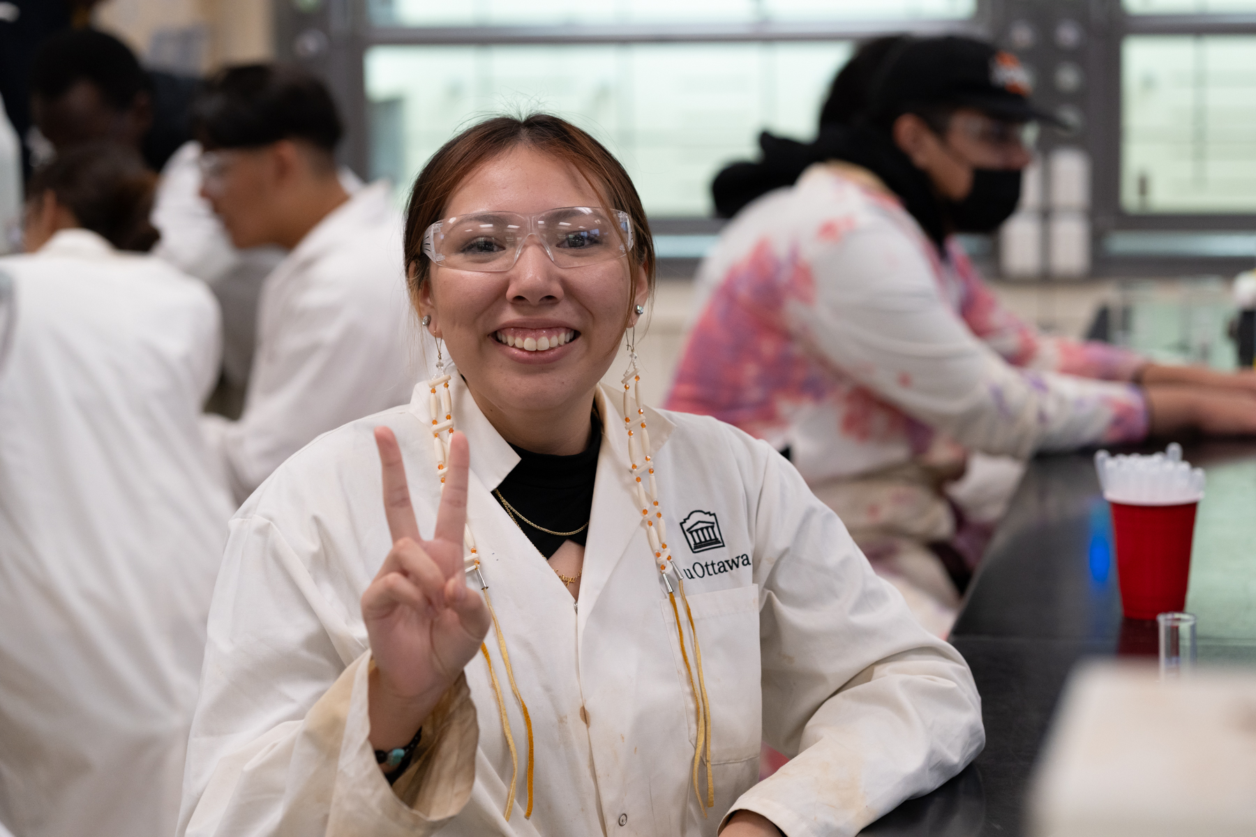 Moose Factory student in white lab coat smiling at the camera and holding up two fingered peace sign