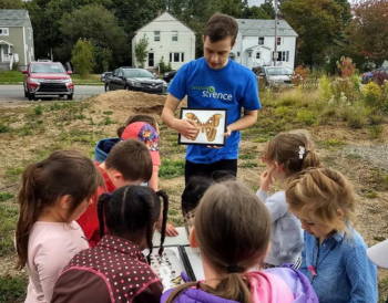  Elementary school students gather outside to look as a volunteer in a blue Let’s Talk Science t-shirt shares a colourful drawing of a butterfly.  