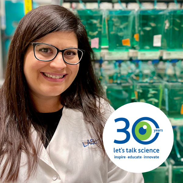 Michelle in a white lab coat smiling at the camera with the Let's Talk Science 30th logo in the corner