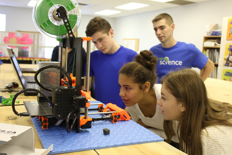 Three students are closely watching a 3D printer in action, as a Let’s Talk Science volunteer looks on. 