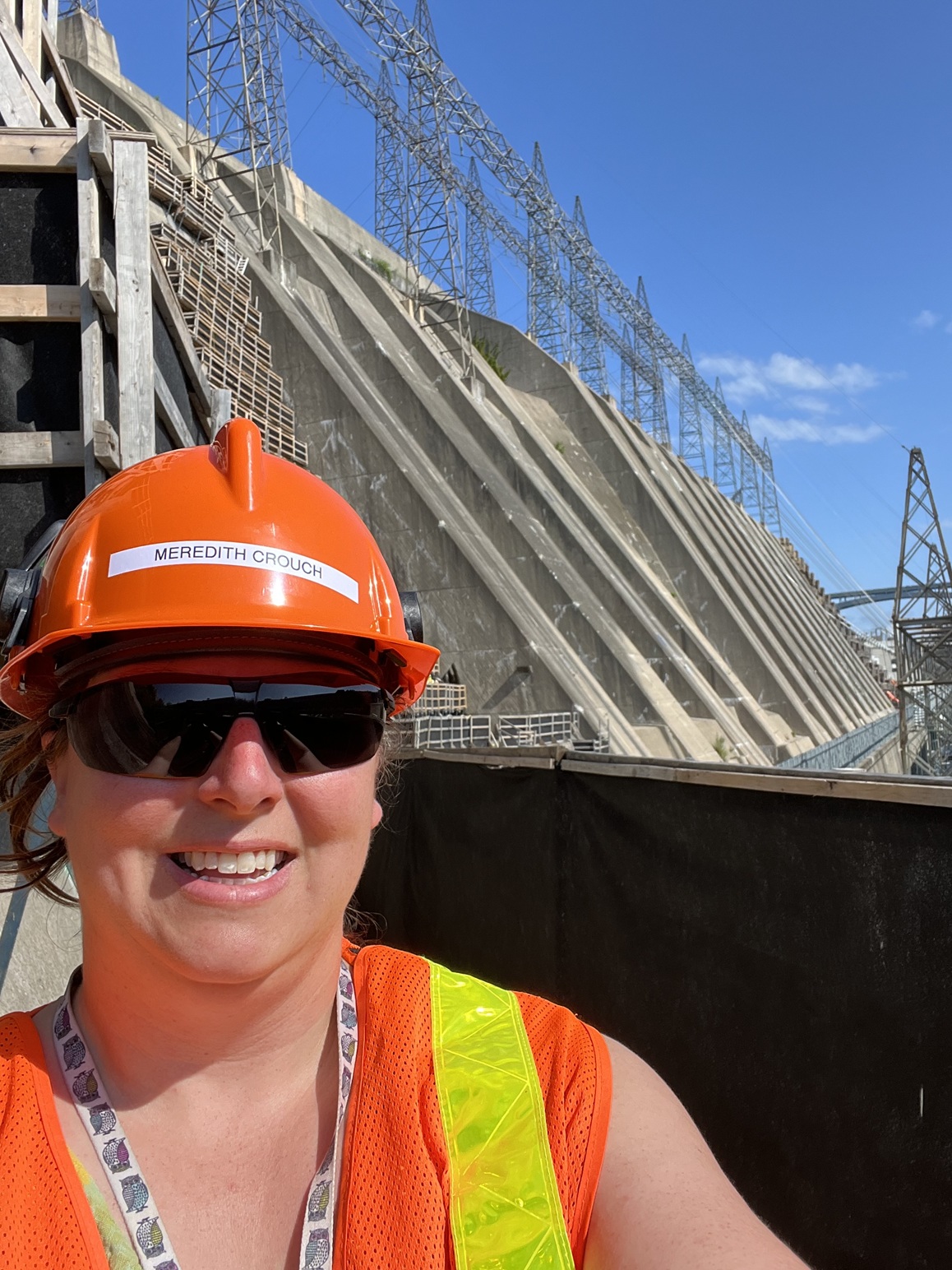 Selfie of Meredith Crouch wearing orange hardhat and orange reflective vest with hydro dam and transmission wires in the background