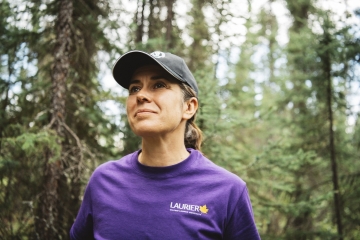 Jennifer Baltzer wearing a bibbed hat, purple tee-shirt with trees in the background.