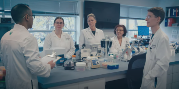 Five people, each wearing a white lab coat, standing around a lab bench that has scientific equipment on it.