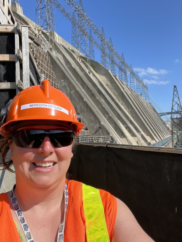 Selfie of Meredith Crouch wearing orange hardhat and orange reflective vest with hydro dam and transmission wires in the background