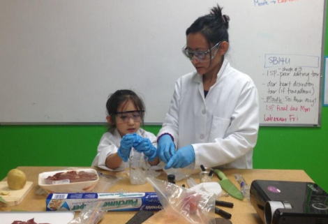 Joelle and her daughter Isabella (pictured at age 5) are standing behind a table as they engage in hands-on classroom activities together. Both are wearing white lab coats with their brown hair tied up, safety goggles and blue plastic gloves. On the table we see a potato, a package of meat, a cutting board, saran wrap, a projector, and miscellaneous plastic bags, markers and utensils spread throughout.