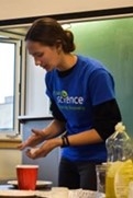 Hannah Richards, a young woman with pale skin and brown hair, demonstrates an activity while wearing her Let's Talk Science blue t-shirt