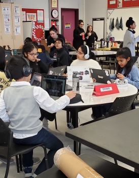 Students sit around a table, each with headphones and a laptop as they complete a simulated space mission together on their screens.