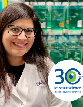 Michelle in a white lab coat smiling at the camera with the Let's Talk Science 30th logo in the corner
