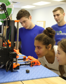 Three students are closely watching a 3D printer in action, as a Let’s Talk Science volunteer looks on. 