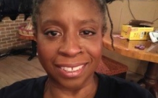 Adrienne Coddett, a Black woman with grey and black locs pulled back from her face, wearing a black tshirt, smiles at the camera