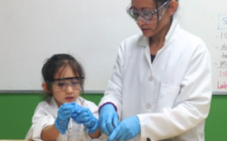 Joelle and her daughter Isabella (pictured at age 5) are standing behind a table as they engage in hands-on classroom activities together. Both are wearing white lab coats with their brown hair tied up, safety goggles and blue plastic gloves. On the table we see a potato, a package of meat, a cutting board, saran wrap, a projector, and miscellaneous plastic bags, markers and utensils spread throughout.