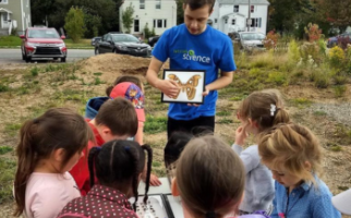  Elementary school students gather outside to look as a volunteer in a blue Let’s Talk Science t-shirt shares a colourful drawing of a butterfly.  
