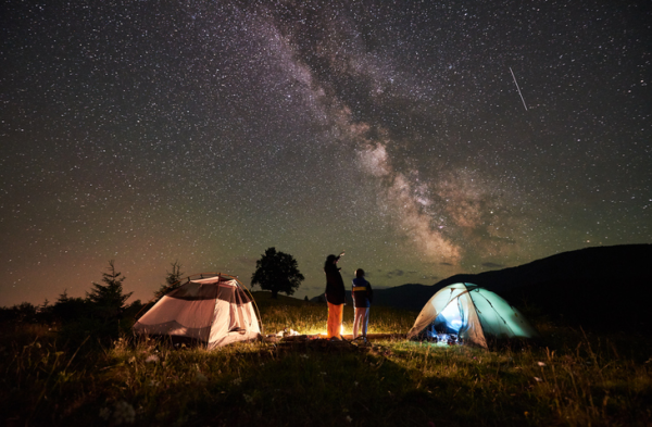 Shown is a colour photograph of a campsite at night, with a streak of bright stars above.
