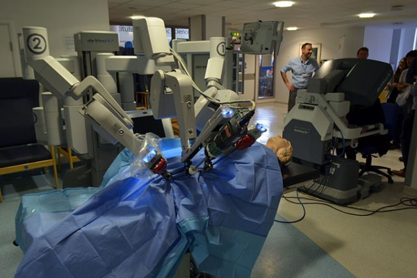 Shown is a colour photograph of three mechanical arms reaching over a model of a patient on a surgical table. 