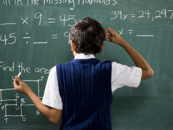 Shown is a colour photograph of a child standing in front of a blackboard, scratching their head. 