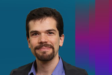 Shown is a colour photograph of a person with short black hair and a beard, wearing a purple shirt and black jacket. They are shown from the shoulders up, on a multicoloured background.
