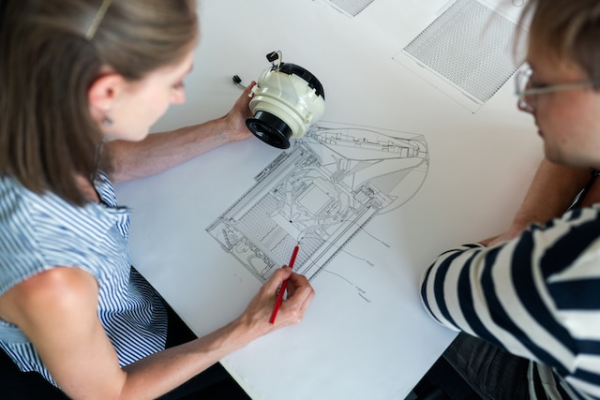 Shown is a colour photograph of two people studying a cylindrical object and a detailed drawing.