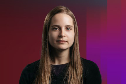 Shown is a colour photograph of a person with long, light brown hair, wearing a black top. They are shown from the shoulders up, on a multicoloured background.